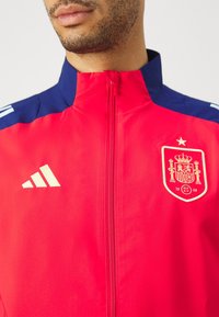 Red zip-up jacket with navy accents, featuring a gold Adidas logo and a gold Spain national emblem on the chest. Smooth texture.