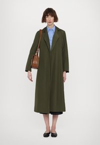 Olijfgroene trenchcoat met een lange silhoët, brede kraag en open voorkant. Gecombineerd met een lichtblauw shirt en een bruine schoudertas. Zwarte flats.