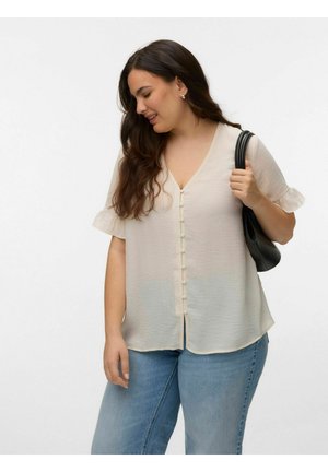 Kobieta o długich brązowych włosach, ubrana w kremową bluzkę na guziki i niebieskie dżinsy, trzyma czarną torebkę na ramię, patrzy w dół i się uśmiecha.