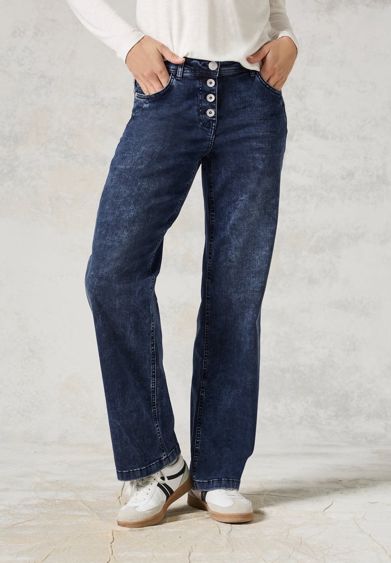 Personne portant un jean bleu foncé taille haute avec braguette à boutons et des baskets blanches, debout sur un sol clair devant un fond neutre.