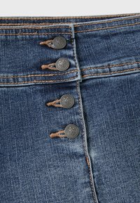 Gros plan sur un jean en denim bleu doté de quatre boutons métalliques, de surpiqûres orange et d'un détail de couture verticale. Aucun logo ou étiquette visible.