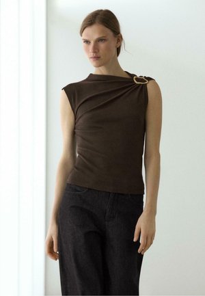 Femme portant un top marron foncé sans manches avec épaule drapée et boucle décorative, associé à un pantalon noir taille haute, debout à l'intérieur.