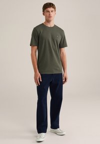 Camiseta verde oliva de manga corta con un pequeño logo, combinada con pantalones azul oscuro y zapatillas blancas. Diseño sencillo, textura de algodón.