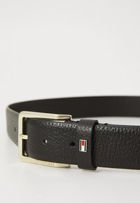 Ceinture noire en cuir texturé avec boucle en or, dotée d'un petit logo rectangulaire rouge, blanc et bleu sur la boucle de ceinture.