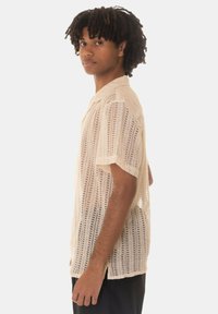 Camicetta a maniche corte in maglia aperta di colore crema. Presenta un colletto e una trama allentata con motivi verticali.