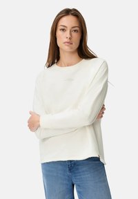 Vrouw met lang bruin haar draagt een off-white sweatshirt en een blauwe spijkerbroek, staande met de armen gekruist tegen een effen achtergrond.