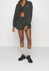 Conjunto deportivo negro que incluye una chaqueta con cremallera, sujetador deportivo y pantalones cortos, complementado con calcetines grises y zapatillas negras con acentos de logo.