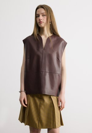 Young woman wearing sleeveless dark brown leather top and yellow plaid pleated skirt, standing against plain white background.