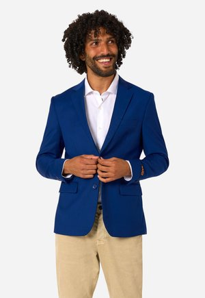 Mann mit lockigem Haar lächelt, knöpft einen königsblauen Blazer über einem weißen Hemd zu, trägt beige Hosen und steht vor einem schlichten Hintergrund.