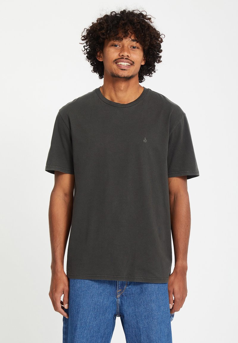 Donkergrijze katoenen t-shirt met een ronde hals en korte mouwen; heeft een klein logo op de borst en een ontspannen pasvorm. Draagt met blauwe spijkerbroek.