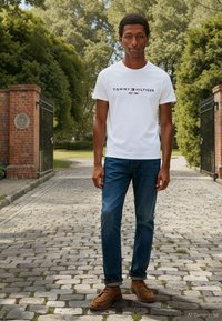 Mees seisab munakiviteel tellistest sambade vahel, tal on seljas valge Tommy Hilfigeri t-särk, sinised teksad ja pruunid seemisnahast kingad välitingimustes.