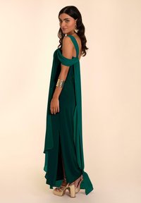 Grünes, bodenlanges Kleid mit drapierten Ärmeln, Seitenschlitzen und einer glatten Textur. Goldene Plateauschuhe und ein metallisches Armband ergänzen den Look.