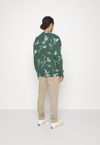 Sweatshirt vert avec motif floral blanc, coupe décontractée, manches longues et poignets côtelés. Associé à un pantalon beige et des baskets claires.