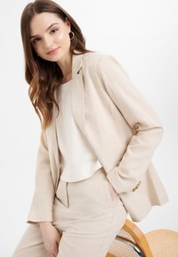 Béžový blazer vyrobený z ľahkého materiálu, s prispôsobeným strihom, klopami a jemným leskom. Nosený cez krémový top, doplnený o zhodné nohavice.