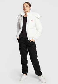Giubbotto imbottito bianco con colletto alto, zip frontale e logo rosso, abbinato a pantaloni della tuta neri e sneaker bianche. Texture complessiva liscia.