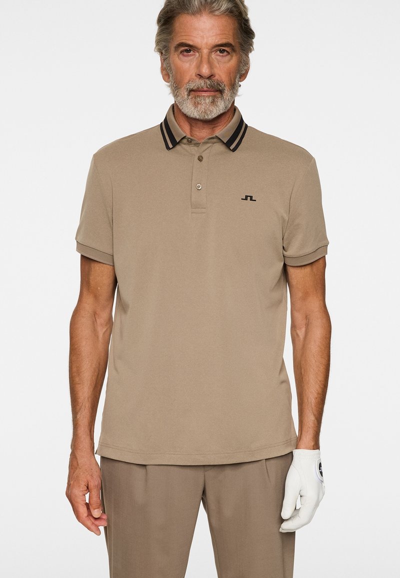 Mężczyzna z siwą brodą ubrany w beżową koszulkę polo, spodnie w tym samym kolorze i białą golfową rękawiczkę na prawej ręce na białym tle.