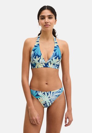 Woman standing wearing blue bikini with green and white large leaf patterns, halter neck top, and matching low-rise bottoms.