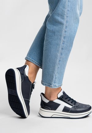 Person trägt schwarz-weiße Sneakers mit dicken Sohlen und hellblauen Jeans, ein Fuß ist leicht angehoben und zeigt die Schuhsohle.