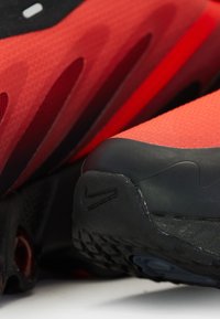 Baskets de sport rouges avec tige en maille texturée, semelle en caoutchouc noire et accent distinctif noir au niveau du talon. Dispose d'ouvertures pour la respirabilité.