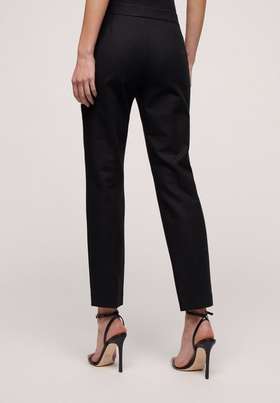 Zwarte taps toelopende broeken met een gladde textuur, voorzien van een discrete tailleband. Gepaard met elegante zwarte hoge hakken met bandjes.