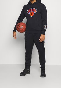 Sweat à capuche noir avec un logo des New York Knicks, assorti à un pantalon de survêtement noir. Tient un ballon de basket ; chaussures de sport noires.