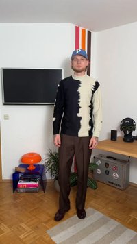 Čierny a krémový vzorovaný sveter s kulatým výstrihom a dlhými rukávmi, v páre s tmavohnedými nohavicami a hnedými topánkami. Na hlave modrá čiapka.
