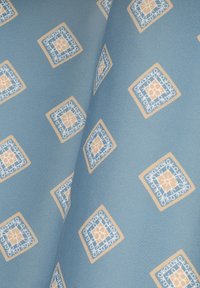 Teksturowana tkanina w jasnym niebieskim kolorze, z geometrycznymi kremowymi i niebieskimi wzorami w kształcie diamentów oraz skomplikowanymi detalami, złożona w delikatne fałdy.