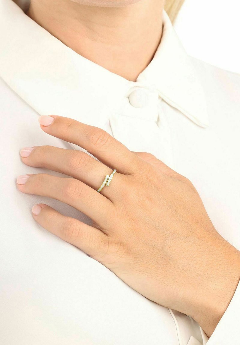 Anillo de oro con una superficie texturizada y un diseño delgado, que presenta un sutil detalle retorcido. La mano descansa sobre una prenda blanca.