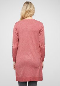Robe tricotée à manches longues, de couleur rouge clair, avec une coupe ample, des poignets côtelés et une ourlet qui tombe sous les hanches, présentant une finition texturée.