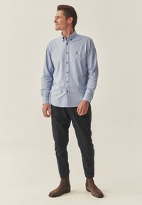 Cămașă călduță, albastră cu nasturi, având un mic logo, nasturi maro și mâneci suflecate, combinată cu pantaloni închiși la culoare și ghete maro până la glezne.