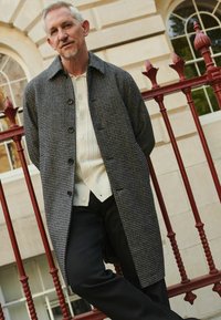 Palton gri cu motive houndstooth, cu închidere cu nasturi, purtat peste un pulover texturat crem, asortat cu pantaloni negri, sprijinit de o balustradă roșie.