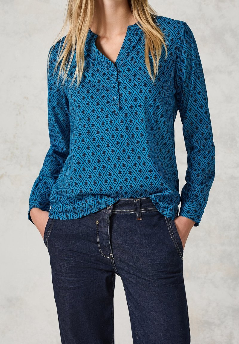 Chemise bleue à motifs avec un col henley, présentant un motif en losange et des manches longues, associée à un jean en denim foncé.