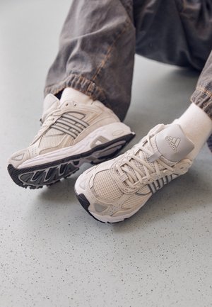 Persoon draagt witte Adidas sneakers met grijze strepen, witte sokken en donkere broek, zittend op een gevlekte vloer.