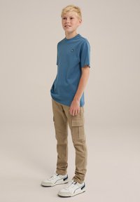 Blå kortärmad t-shirt med rund halsringning och liten logotyp, ihop med beige cargobyxor med flera fickor, och vita sneakers med svarta detaljer.