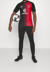 Camiseta de fútbol negra y roja con rayas verticales, combinada con pantalones negros y botas negras. El jugador sostiene un balón de fútbol en blanco y negro.