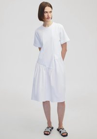 Witte jurk met korte mouwen, met een aansluitende top en geplooide rok. Gemaakt van gladde stof, gecombineerd met zwart-witte sandalen met een patroon.