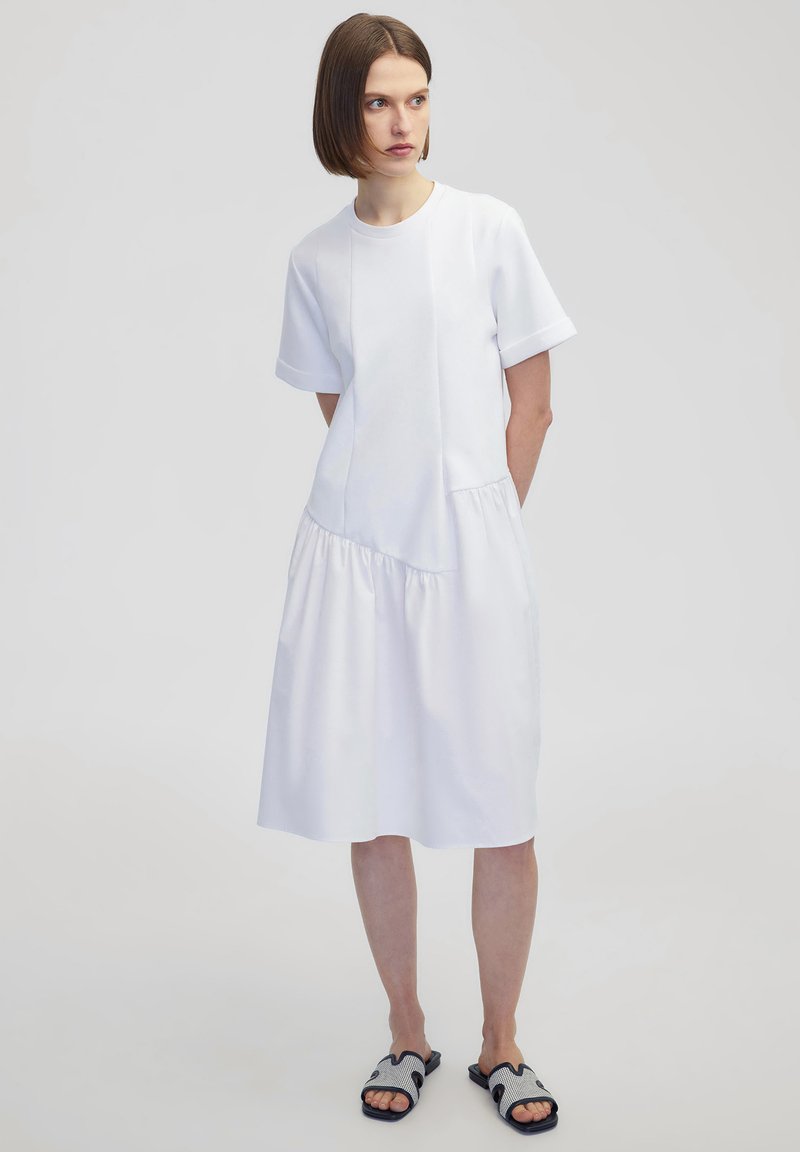Witte jurk met korte mouwen, met een aansluitende top en geplooide rok. Gemaakt van gladde stof, gecombineerd met zwart-witte sandalen met een patroon.