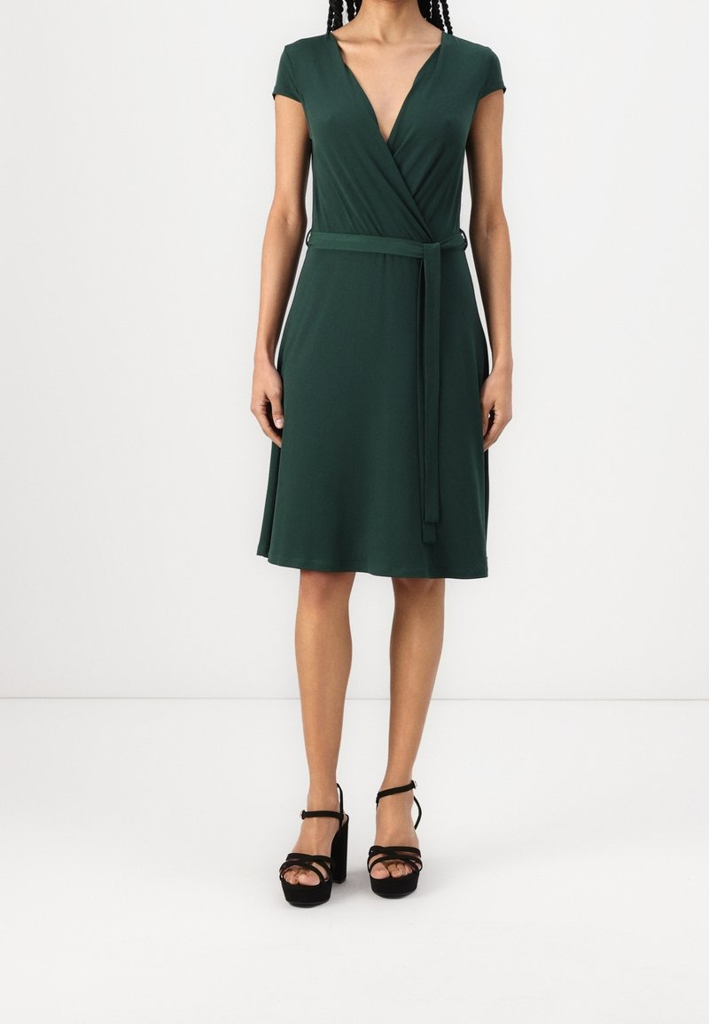 Femme portant une robe portefeuille vert foncé, longueur genoux, manches courtes, avec une ceinture assortie et des talons compensés noirs.
