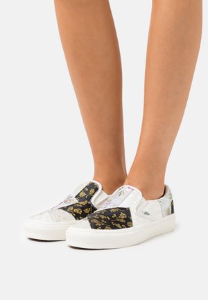 Et par hvide slip-on Vans sko med sort og guld blomsterpatchwork, der bæres på bare fødder mod en hvid baggrund.