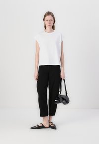 Bílý krátký rukáv top, černé volné kalhoty a černé ploché boty. Drží malou černou kabelku z lakované kůže. Minimalistický design.