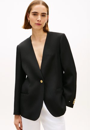 Vrouw met kort bruin haar, gekleed in een zwarte blazer met gouden knopen en witte broek, staand tegen een effen lichte achtergrond.