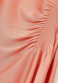 Tessuto color pesca con una texture liscia e una leggera lucentezza. Presenta dettagli arricciati su un lato con accentuazioni di smock elasticizzato.