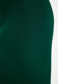 Material de tricot verde cu nervuri, cu așezare verticală, textură netedă și formă ajustată, evidențiind o construcție fără cusături pe latură.