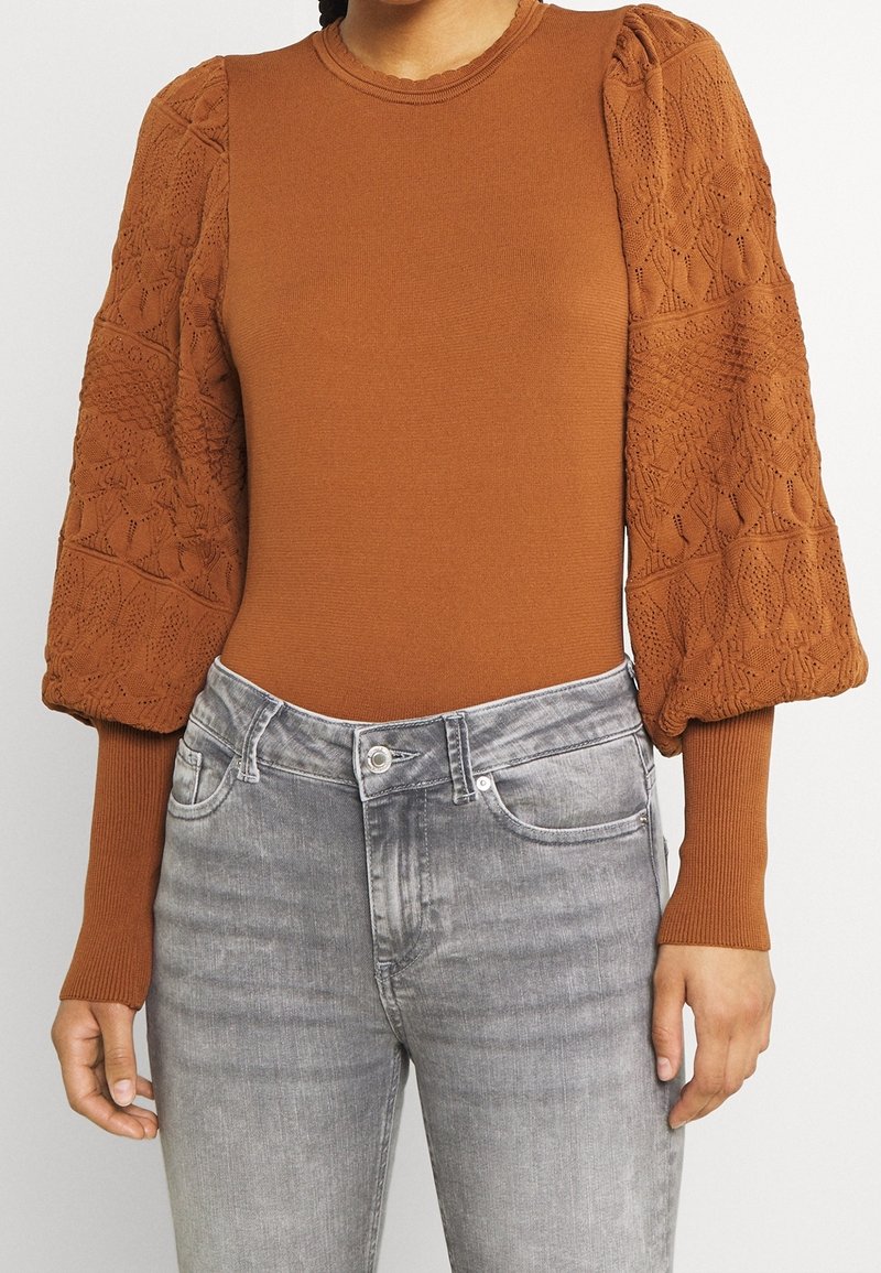 Haut en maille couleur rouille avec des manches en dentelle texturée et volumineuse, associé à un jean gris délavé taille haute.