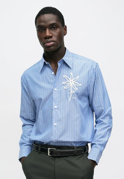 Jauns vīrietis, valkājot zilu un baltu svītrainu kreklu ar pogām, uz kura ir izšūta balta zieda dizains, melnu jostu un tumšas bikses.