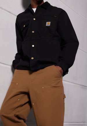 Personne portant une veste Carhartt noire boutonnée et un pantalon de travail marron, debout contre un mur blanc texturé avec une main dans la poche.