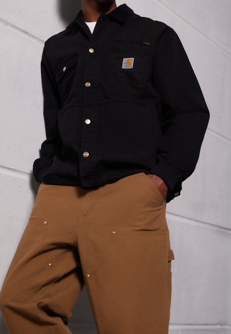Personne portant une veste Carhartt noire boutonnée et un pantalon de travail marron, debout contre un mur blanc texturé avec une main dans la poche.