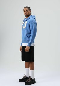 Blauwe hoodie met crème nummer "23", zakken aan de voorkant, gedragen over een wit shirt, gecombineerd met zwarte korte broek en zwarte schoenen met roze veters.