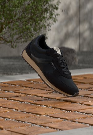 Czarny sneaker GANT z beżową podeszwą unoszący się nad wzorzystą kostką brukową na zewnątrz, w tle zielone krzewy.