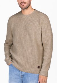 Beige gerippter Pullover mit langen Ärmeln, Rundhalsausschnitt und strukturiertem Finish. Kleines braunes Logo-Patch auf der unteren linken Seite.